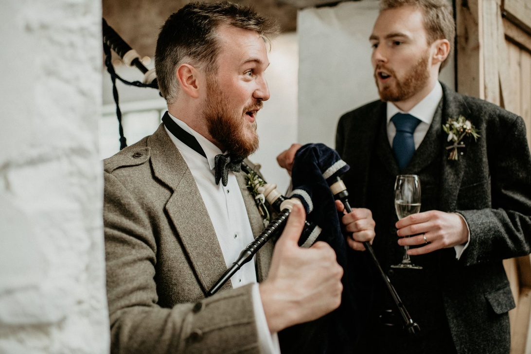 Två män i tweedkostymer håller tal under ett bröllop, en talar animerat med mikrofon och champagneglas.