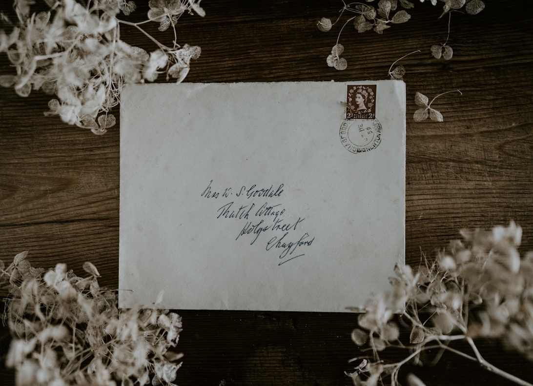 An antique envelope with handwritten address and vintage stamp surrounded by dried flowers on dark wood, evoking nostalgic wedding correspondence.